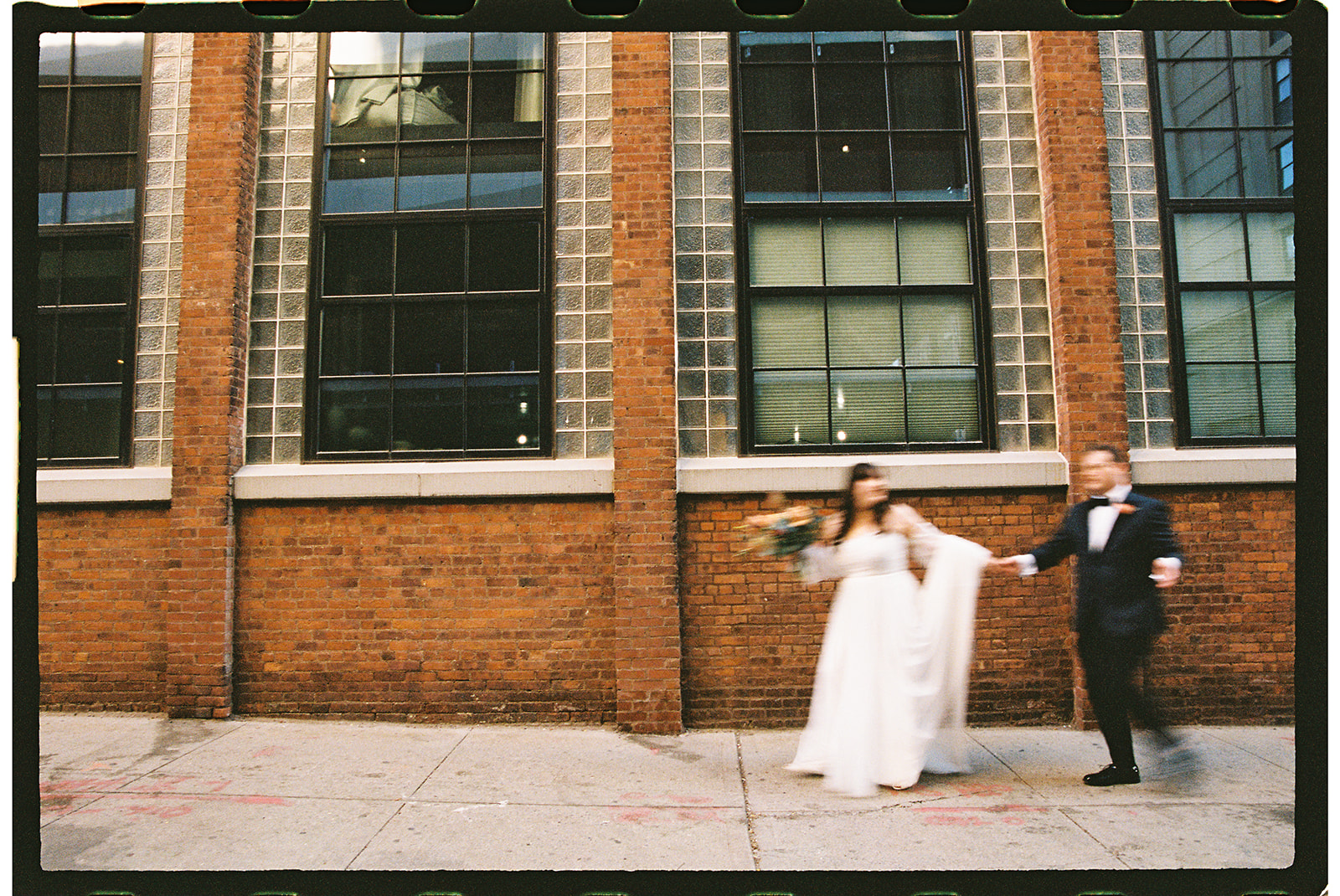 blurry film photo of couple walking down the street in front of a brick building in DUMBO brooklyn new york